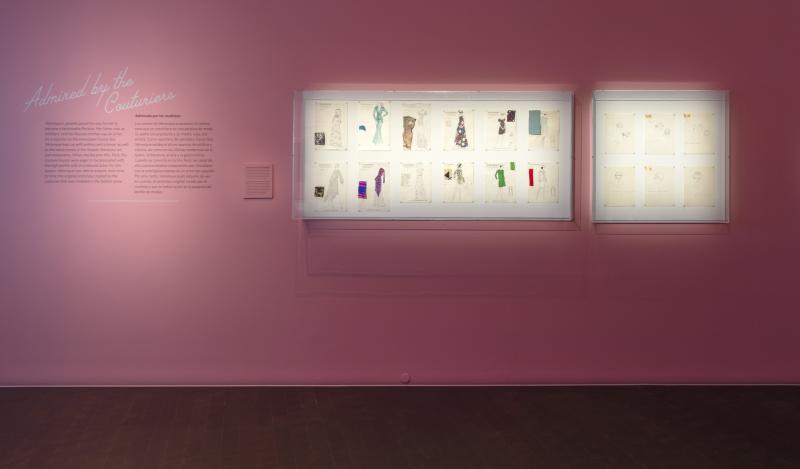 Photograph of part of the exhibition that features eighteen different fashion sketches hung on a pink wall.