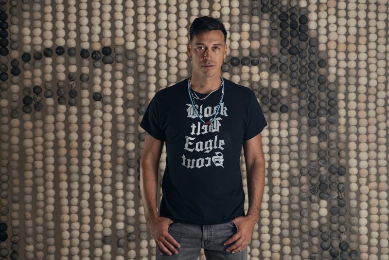 Color photograph of the artist, Cannupa Hanska Luger, wearing a black tee shirt with white text, beaded necklaces, and jeans standing in front of his artwork.