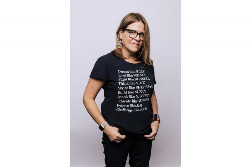 Color photograph of the artist, Marie Watt, wearing a black tee shirt with white text and black jeans.