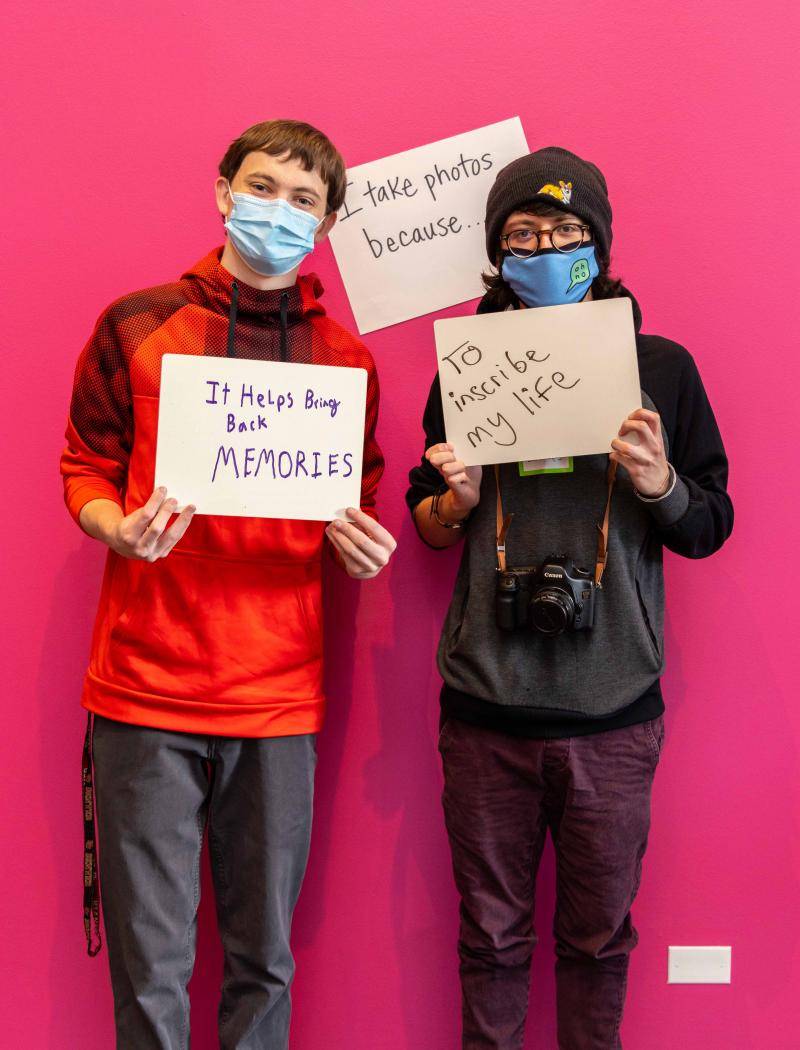 Two teens posing in the exhibition with written signs explaining their reasons why they take photos