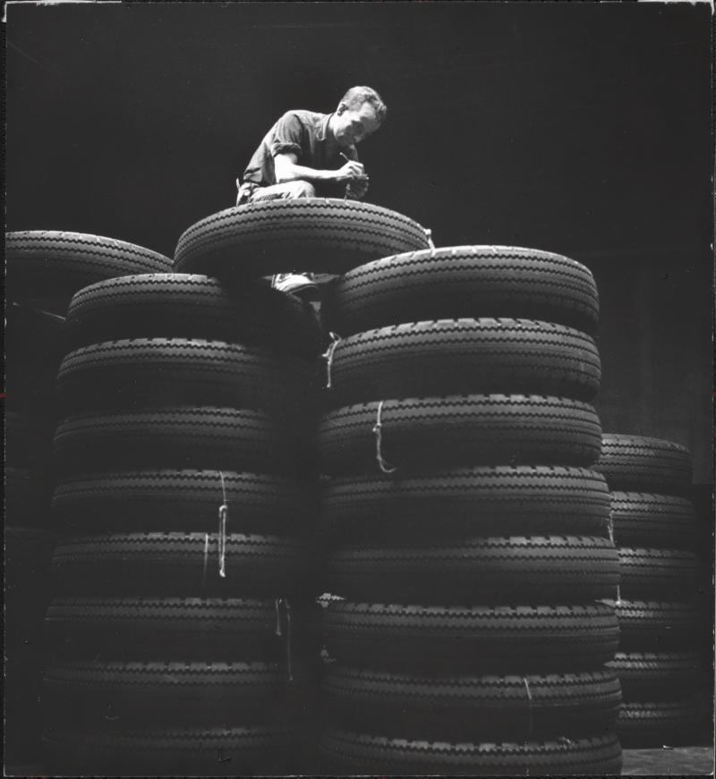 Black and white photograph of a man atop a mound of car tires