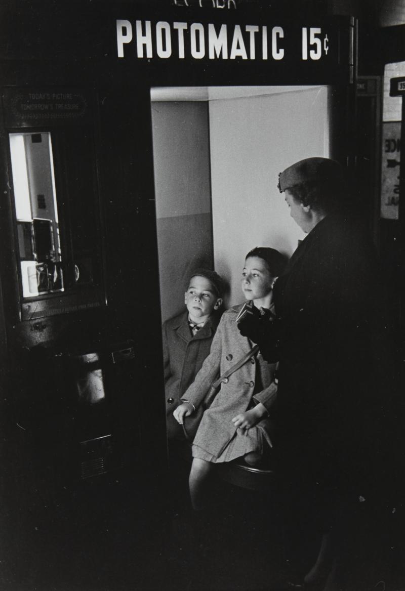 Black and white photograph of two young boys in a photobooth, overlooked by their mother