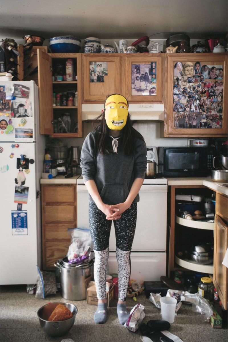 Young woman standing in messy kitchen wearing a yellow mask over her face