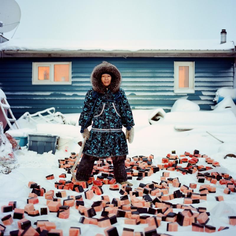 Photograph of an Inuit woman dressed in winter gear in front of her home in snowy Alaska
