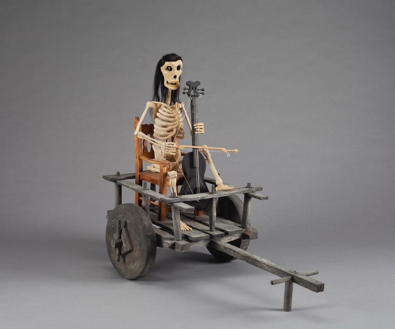 Sculpture of a skeletal man holding an instrument while riding in a wooden cart
